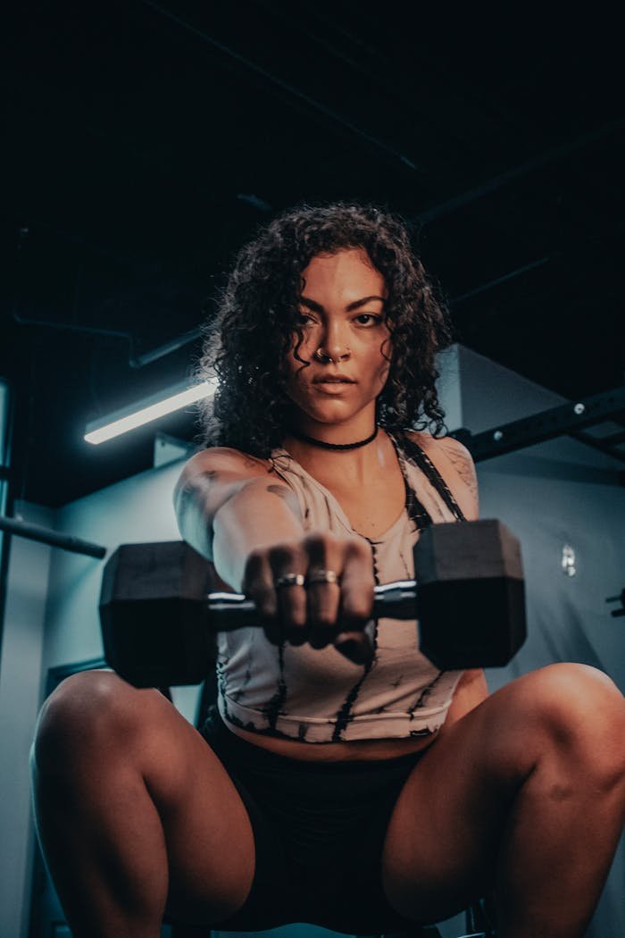 Focused woman with curly hair lifting a dumbbell in a dimly lit gym, showcasing strength.