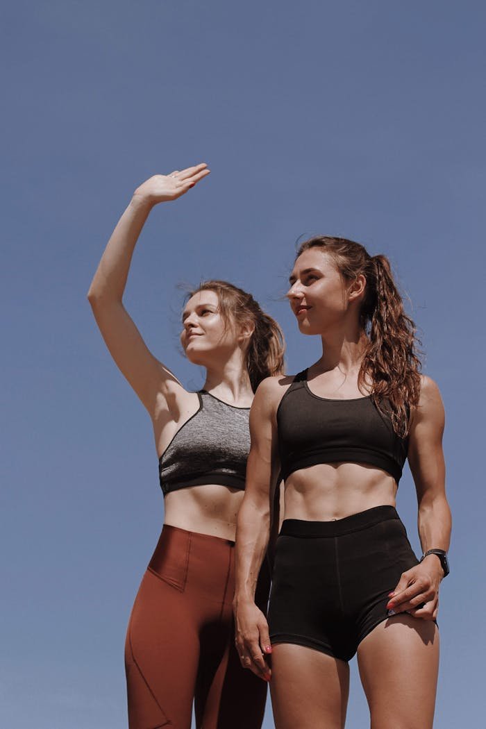 Two women in athletic wear pose confidently outdoors against a clear blue sky.